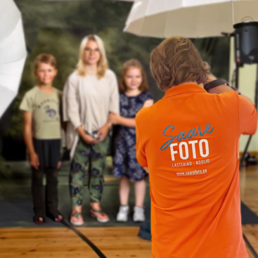 Esiplaanil on fotograaf, seljas Saarefoto oranž särk ning seal peal Saarefoto logo, lapsed naeratavad ja poseerivad fotograafile.